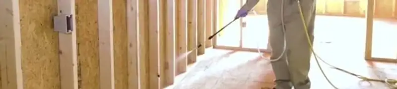 Termite treatment technician applying spray along framing and unfinished walls in Oakland, CA
